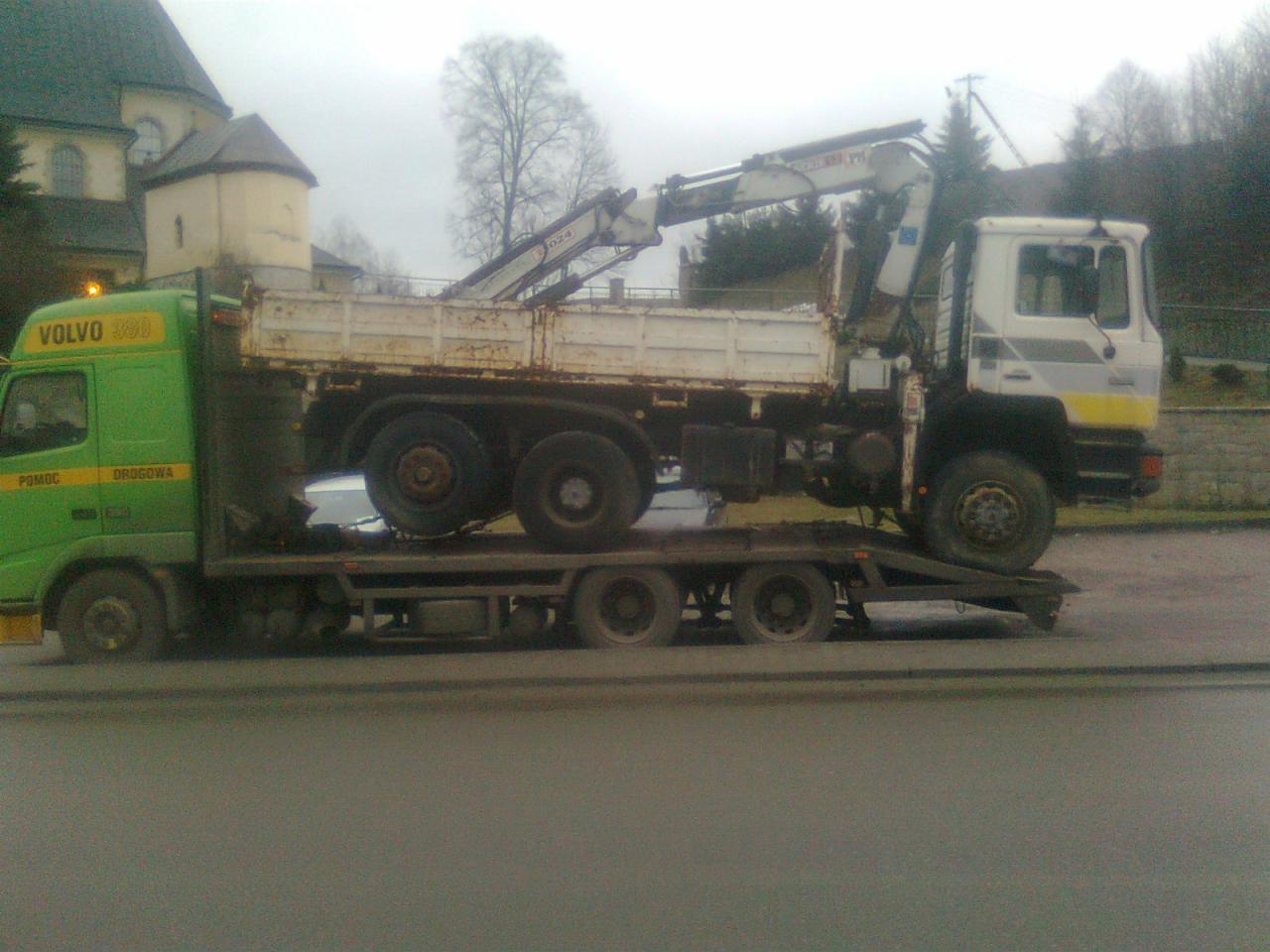 Laweta przewozi ciężarówkę z dźwigiem na tle zabudowań. Zielona kabina Volvo 360. Specjalistyczny transport drogowy z wykorzystaniem niskopodwoziowej platformy.