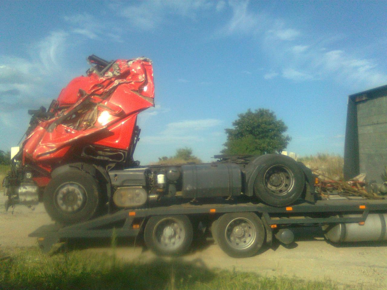 Laweta z uszkodzoną, czerwoną kabiną ciężarówki po wypadku, na tle błękitnego nieba i zieleni. Widoczne zniekształcenia karoserii.