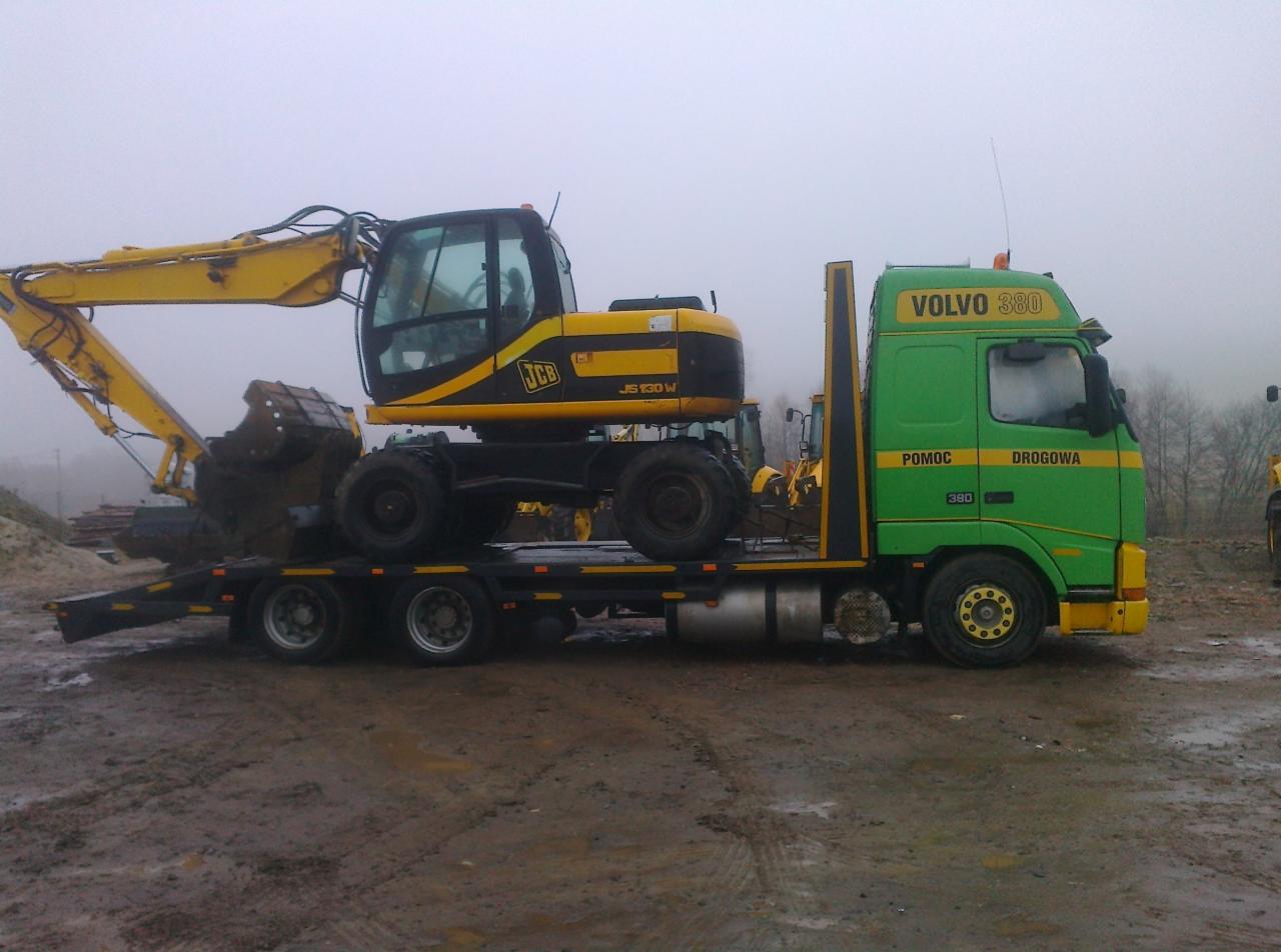 Żółta koparka JCB JS130W przewożona na niskopodwoziowej lawecie Volvo 380 Pomoc Drogowa w pochmurny dzień. Widoczny fragment terenu budowy.