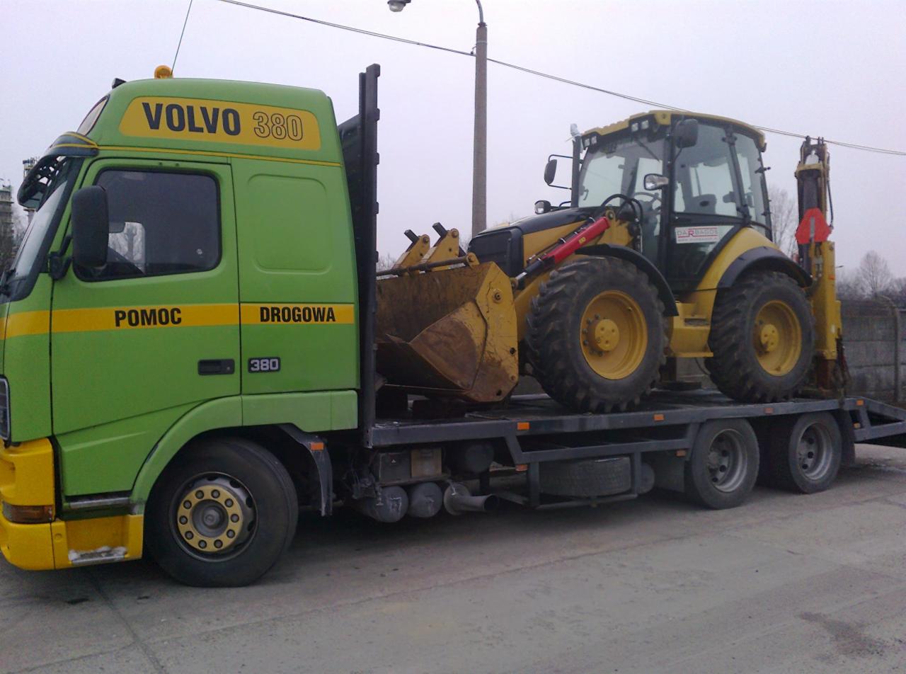 Zielona laweta Volvo 380 z żółtym traktorem z przodu. Pomoc drogowa. Transport maszyny budowlanej na niskopodwoziowej platformie. Szare niebo w tle.