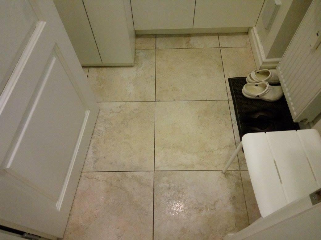 Beżowe płytki podłogowe w łazience z białymi szafkami, drzwiami i taboretem. Na wycieraczce leżą białe klapki. Widok z góry, oświetlenie sztuczne.