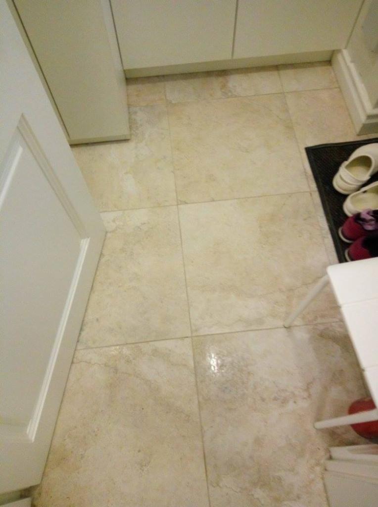 Jasne, kwadratowe płytki podłogowe z delikatnym wzorem w łazience, widoczne buty na wycieraczce i fragment białych mebli.