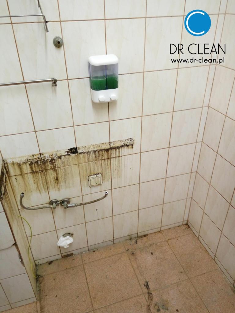 Zanieczyszczona łazienka z widocznymi śladami brudu na ścianach, podłodze i armaturze. Dozownik z płynem i logo firmy sprzątającej w górnym rogu.