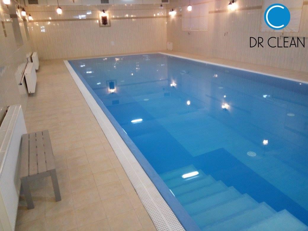 Błękitny basen kryty z schodami, płytkami wykończony, obok ławka i grzejniki. Logo firmy sprzątającej w rogu.