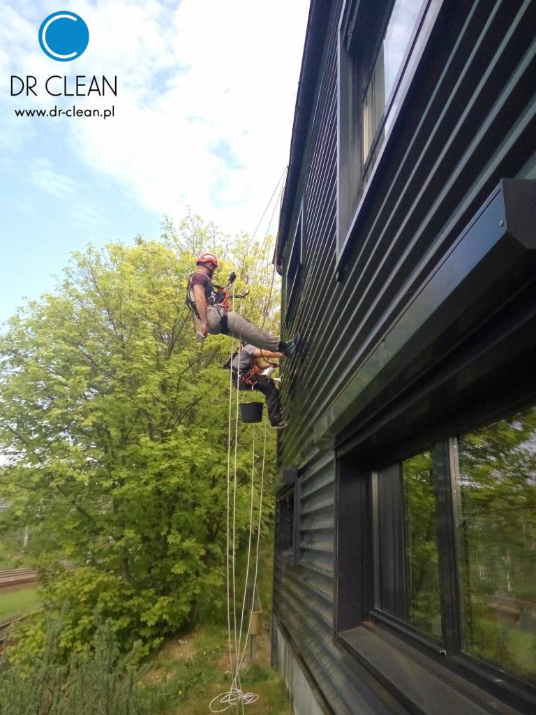 Alpinista myje okna na wysokości w nowoczesnym budynku z ciemną elewacją. Widoczne liny, uprząż i wiadro. Duże okna odbijają zieleń drzew.