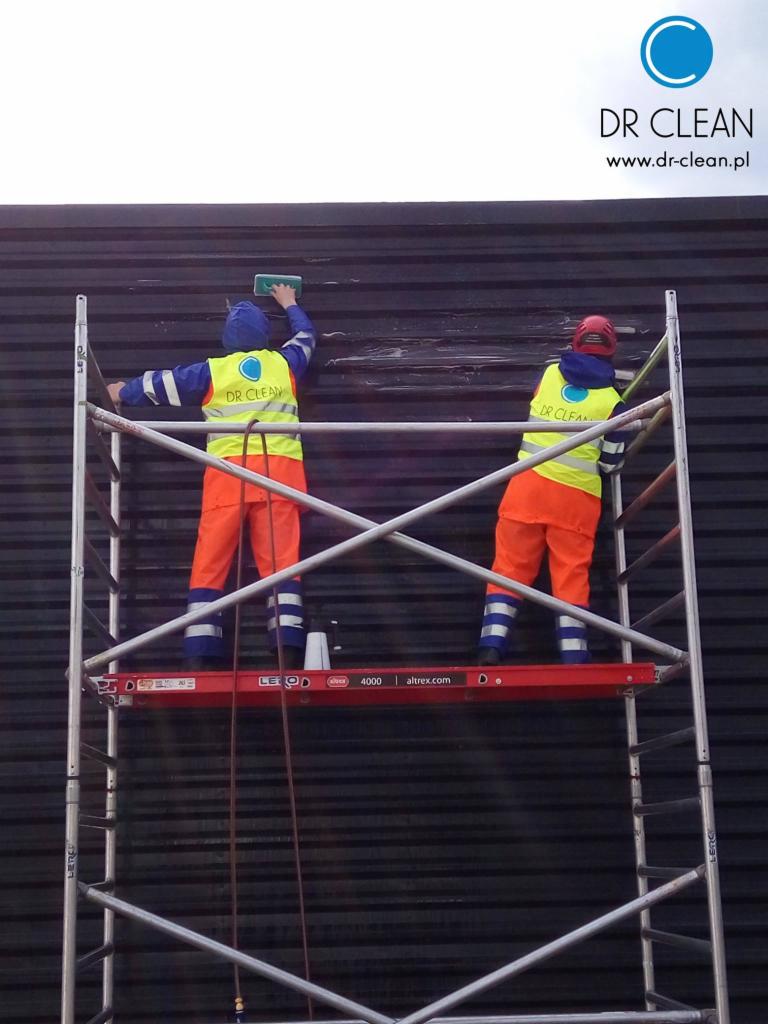 Dwóch pracowników na rusztowaniu czyści ciemną, żłobkowaną elewację budynku. Używają gąbek i noszą odblaskowe kamizelki z logo firmy.
