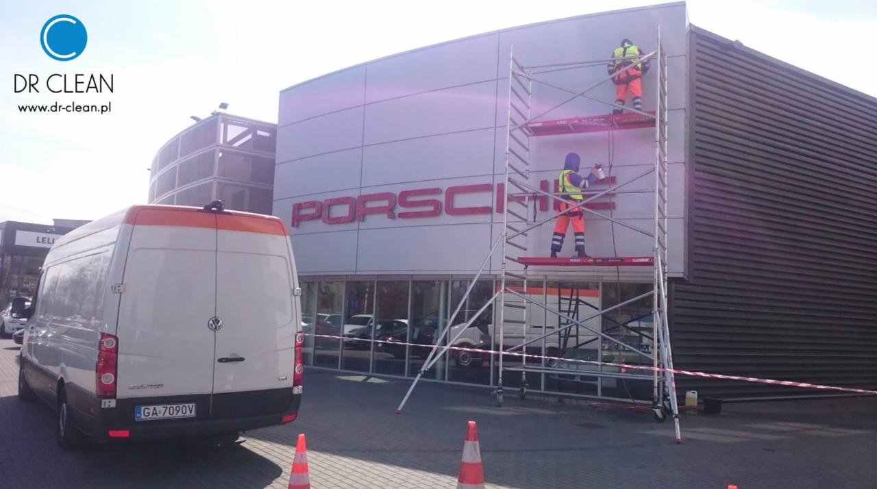Ekipa w jaskrawych strojach na rusztowaniu czyści elewację budynku Porsche. Biały van z logo firmy sprzątającej stoi obok, a pomarańczowe pachołki wyznaczają strefę roboczą.