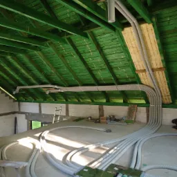 Wnętrze w trakcie instalacji systemu wentylacji z widocznymi rurami wentylacyjnymi, zieloną drewnianą konstrukcją dachu i oknem w tle.