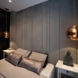 Nowoczesna sypialnia z szarym panelem ściennym, miedzianymi lampami wiszącymi i stolikiem nocnym z figurką Buddy.