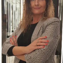 Portret kobiety w szarej marynarce z założonymi rękami, z pomarańczowym manicure i złotymi pierścionkami, na tle ściany z pionowymi, podświetlanymi elementami.