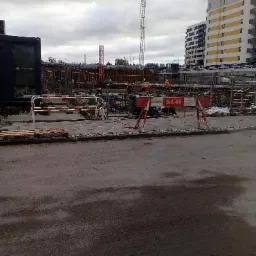 Plac budowy w Poznaniu z widocznym żurawiem, konstrukcjami zbrojeniowymi i pracownikami w pomarańczowych kamizelkach, zabezpieczony barierkami. W tle widać budynek mieszkalny w trakcie budowy.