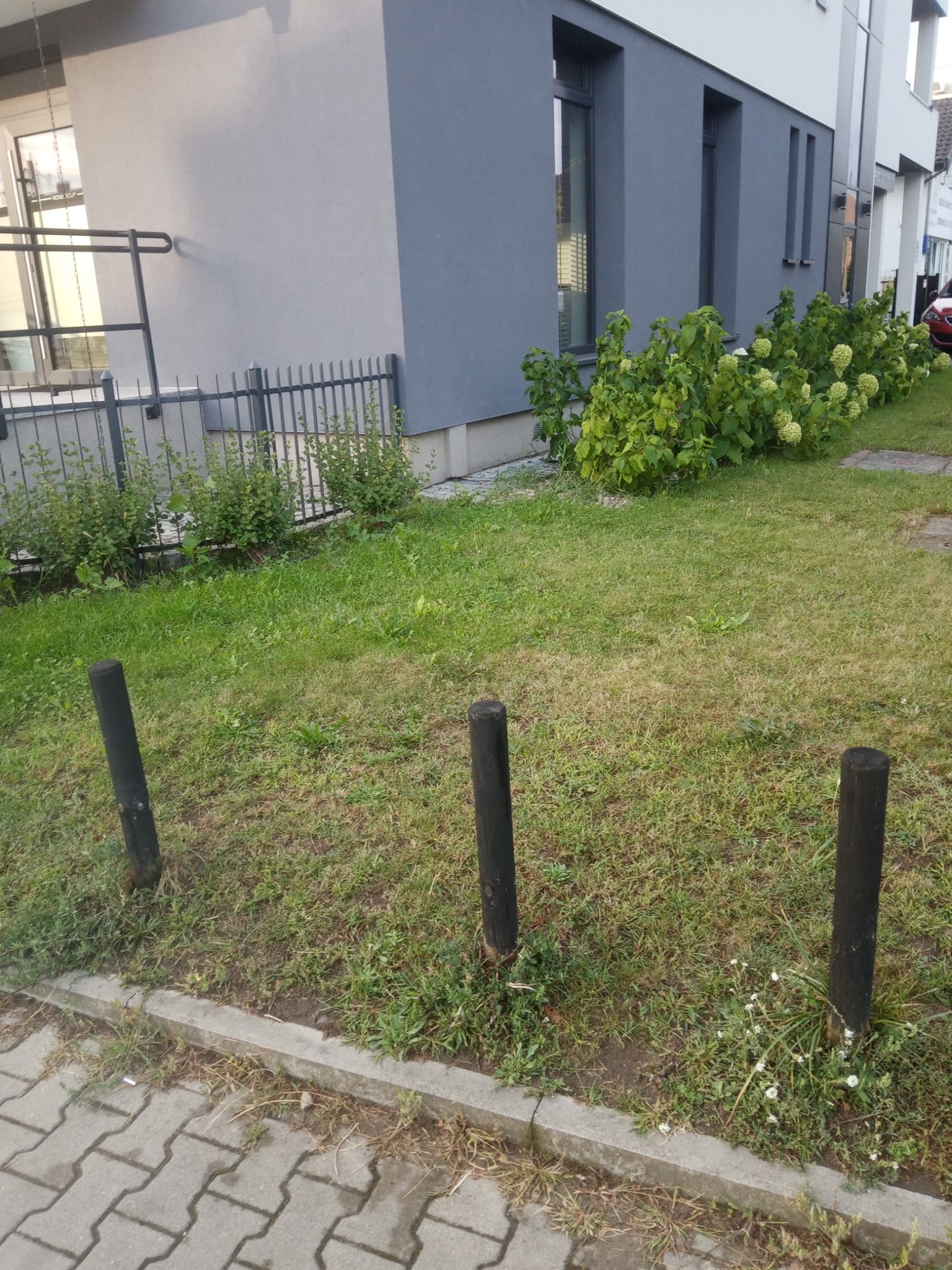 Fragment elewacji dwukondygnacyjnego budynku z szarą fasadą, ogrodzeniem i zielenią, widoczne trzy drewniane słupki odgradzające chodnik od trawnika.