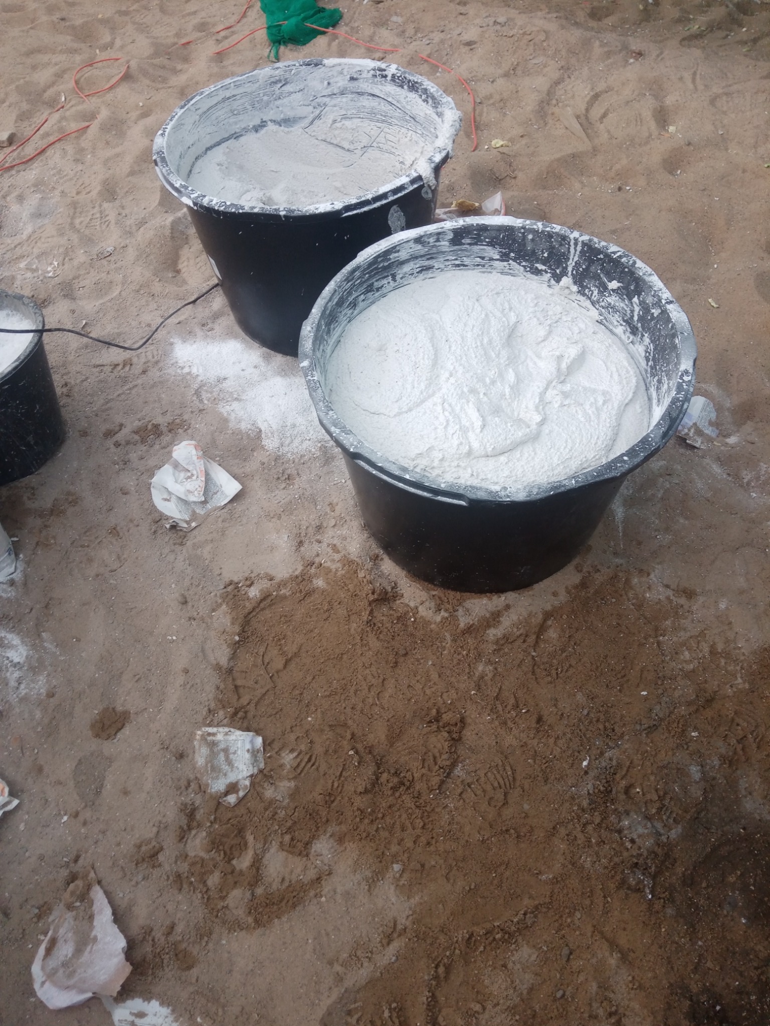 Dwa czarne wiadra wypełnione zaprawą murarską na piaszczystym podłożu z widocznymi śladami butów i fragmentami opakowań po materiałach budowlanych.