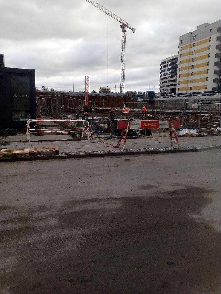 Plac budowy w Poznaniu z widocznym żurawiem, konstrukcjami zbrojeniowymi i pracownikami w pomarańczowych kamizelkach, zabezpieczony barierkami. W tle widać budynek mieszkalny w trakcie budowy.