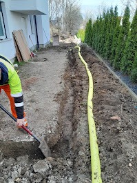 Wykopany wąski rów w ziemi wzdłuż ogrodzenia z tui, żółta rura ułożona obok wykopu, pracownik z łopatą na pierwszym planie, budynek w tle.