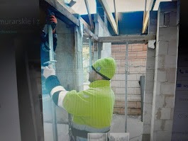 Pracownik w zielonej kurtce i czapce podtrzymuje metalową podporą drewniany element konstrukcji stropu na budowie domu z białych bloczków.