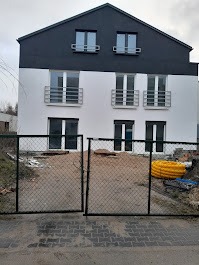 Dwukondygnacyjny dom z białymi ścianami i czarnym dachem, widoczny przez otwartą bramę z siatki. Na podwórku widoczne materiały budowlane i żółta rura karbowana. Pochmurne niebo w tle.