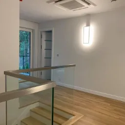 Wnętrze domu z drewnianą podłogą, szklaną balustradą schodów i kasetonową jednostką klimatyzacyjną sufitową w białej obudowie