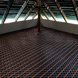 Pomarańczowe rury ogrzewania podłogowego ułożone na czarnej macie izolacyjnej w surowym wnętrzu poddasza z widoczną konstrukcją dachu.