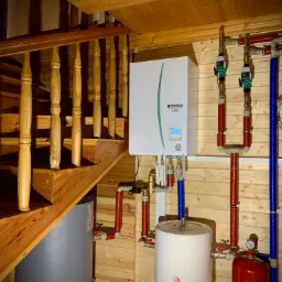 Wnętrze domu z drewnianym wykończeniem: zainstalowana pompa ciepła Ecodan obok schodów, z widocznymi rurami w izolacji termicznej i zbiornikiem buforowym.