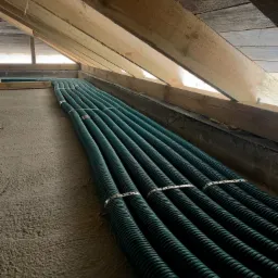 Zielone, karbowane rury wentylacyjne ułożone równolegle na betonowej posadzce pod drewnianą konstrukcją dachu, zabezpieczone metalowymi opaskami.