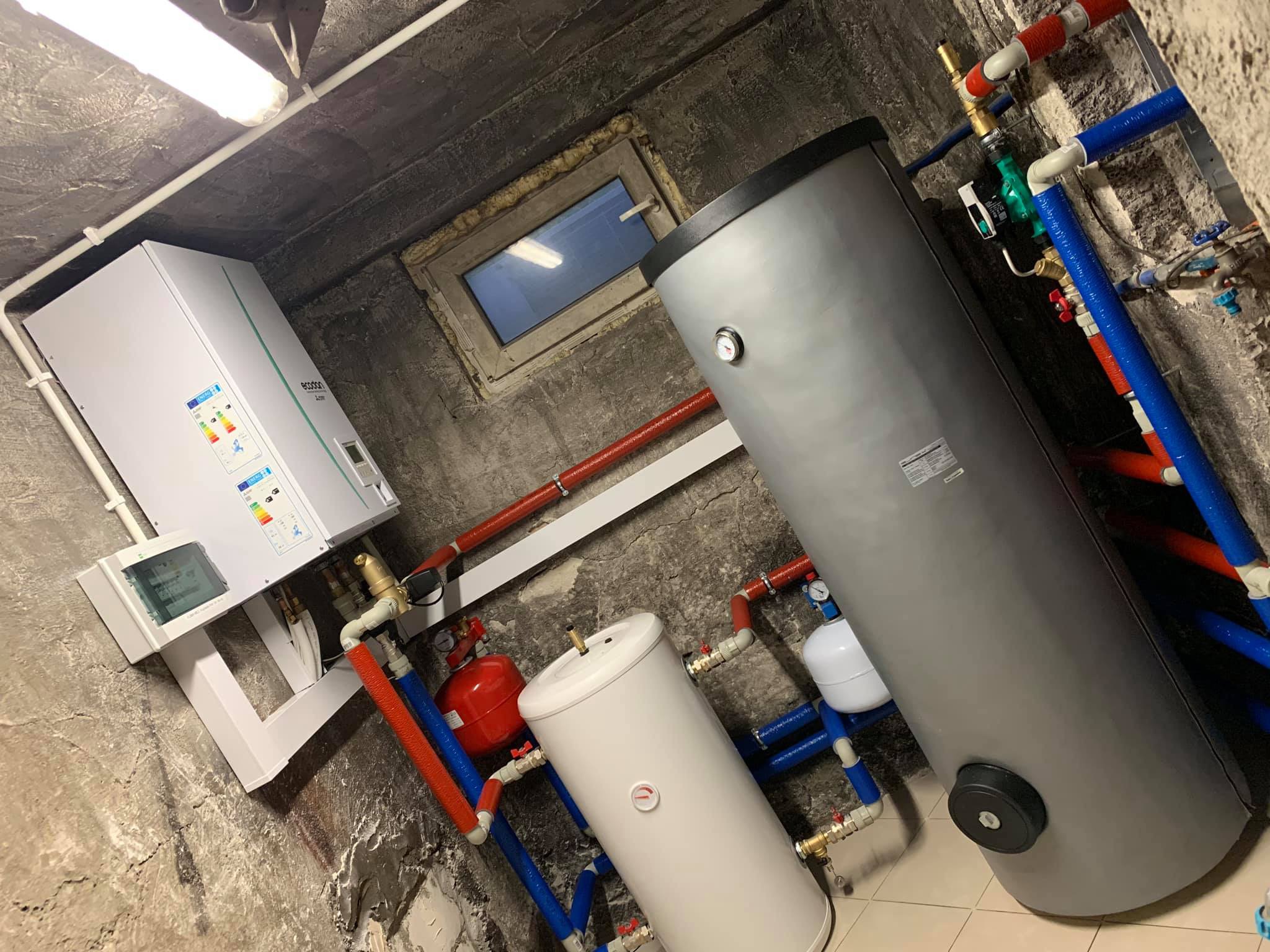 Zestaw instalacji grzewczej w piwnicy z szarym zbiornikiem buforowym, białym bojlerem i kotłem gazowym na ścianie z widocznymi rurami w izolacji czerwonej i niebieskiej.