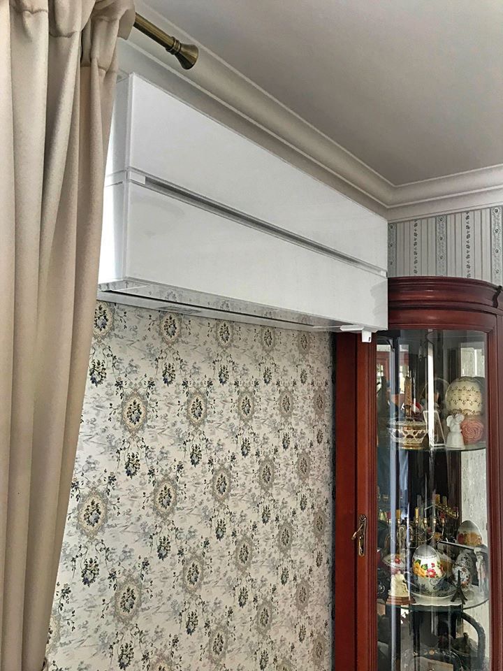 Biała jednostka wewnętrzna klimatyzacji zamontowana pod sufitem w pomieszczeniu z tapetą w kwiaty i zasłonami.