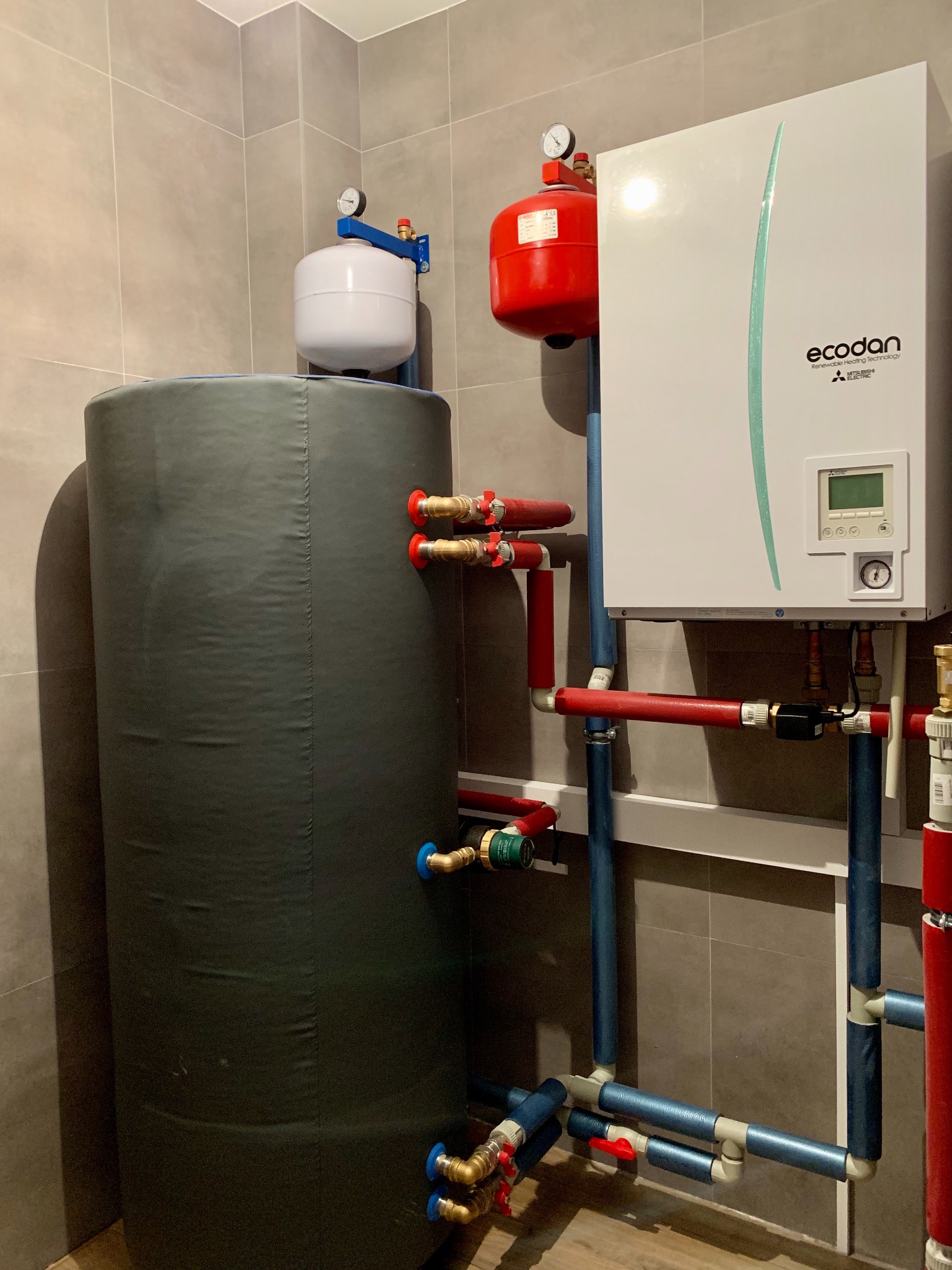 Zestaw urządzeń grzewczych w kotłowni: pionowy zbiornik akumulacyjny w szarej otulinie, biała jednostka sterująca Ecodan Mitsubishi Electric, czerwony zbiornik wyrównawczy, instalacja z rur...