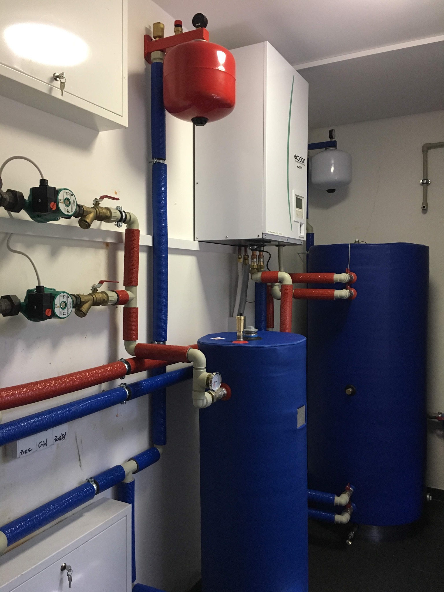 Zakończona instalacja kotła grzewczego z zasobnikami wody, rurami w izolacji i pompami obiegowymi w Starych Kupiskach. Widoczny biały kocioł marki Ecodan.