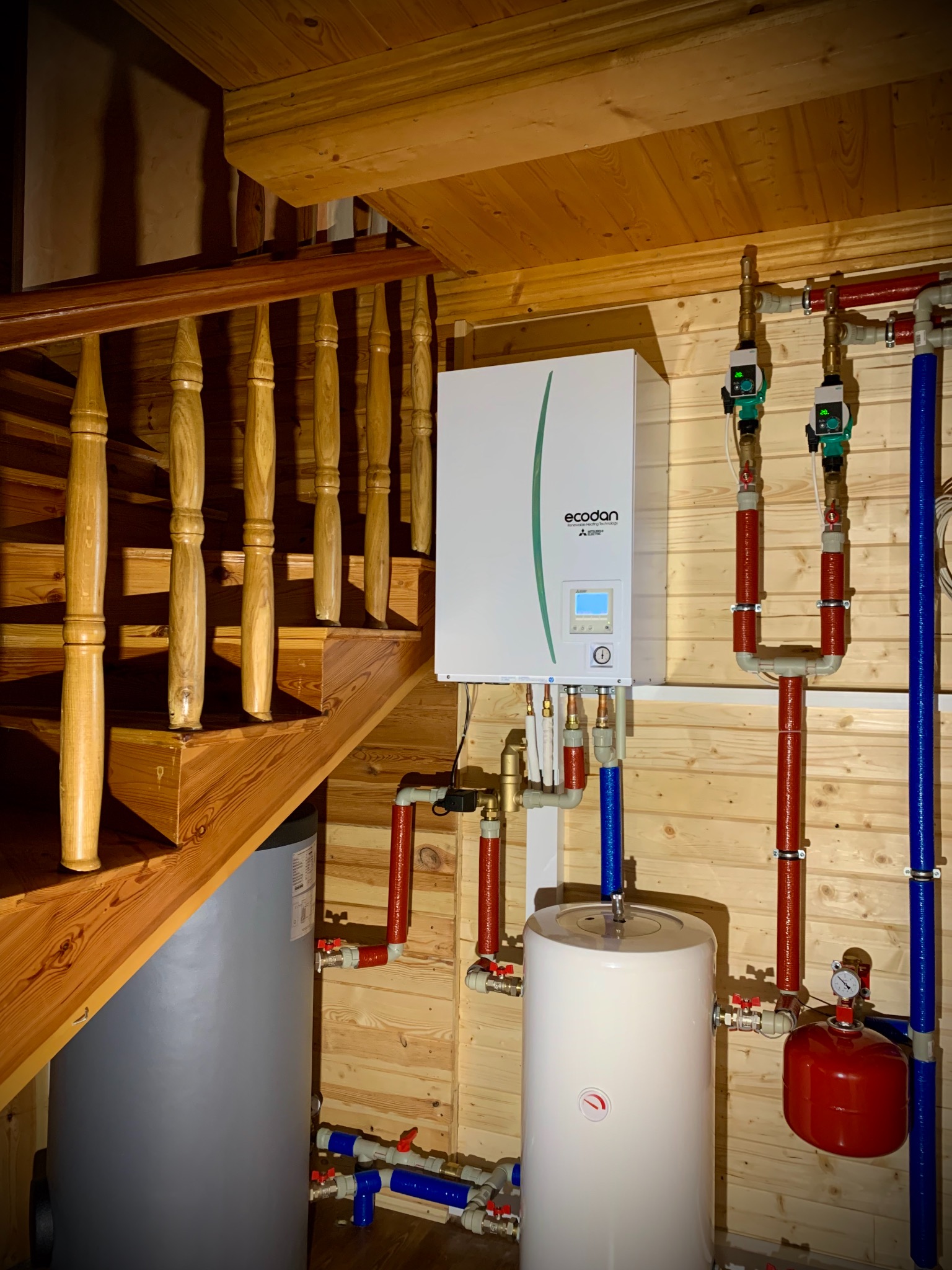 Wnętrze domu z drewnianym wykończeniem: zainstalowana pompa ciepła Ecodan obok schodów, z widocznymi rurami w izolacji termicznej i zbiornikiem buforowym.