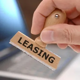 Dłoń trzyma drewniany stempel z napisem LEASING na tle rozmytego laptopa. Stempel skierowany w stronę widza, oświetlenie studyjne.