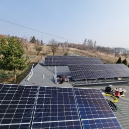 PROENERGY KAROL MIŚKIEWICZ - Montaż paneli słonecznych na dachu domu jednorodzinnego, widok z góry, instalator w trakcie pracy, zwój kabli i narzędzia obok.