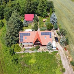 PROENERGY KAROL MIŚKIEWICZ - Zdjęcie z lotu ptaka przedstawia dom z czerwonym dachem, na którym zamontowano panele fotowoltaiczne. Obok domu zaparkowane są dwa samochody, a całość otoczona jest zielenią.