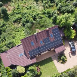 PROENERGY KAROL MIŚKIEWICZ - Widok z lotu ptaka na dom z brązowym dachem i panelami słonecznymi, otoczony bujną zielenią i dwoma samochodami na podjeździe z kostki.