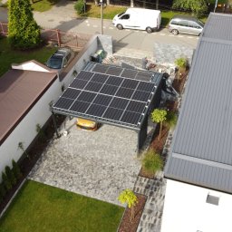 PROENERGY KAROL MIŚKIEWICZ - Widok z góry na carport z panelami słonecznymi, z żółtym samochodem pod spodem, otoczony kostką brukową i zielenią, w tle domy jednorodzinne i samochody na ulicy.