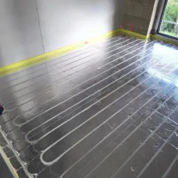 Rozłożona instalacja ogrzewania podłogowego na folii aluminiowej w surowym pomieszczeniu z oknem, widoczne rury w kształcie pętli i żółta taśma przy ścianach.