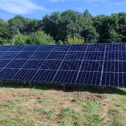 ProEnergy - Panel fotowoltaiczny ustawiony na metalowych wspornikach, widoczny na tle zieleni drzew i błękitnego nieba, z ziemią częściowo pokrytą trawą i śladami prac ziemnych.