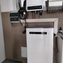 PROENERGY KAROL MIŚKIEWICZ - Biały magazyn energii GoodWe z podłączonymi kablami i ładowarką, zamontowany na ścianie z beżowych płytek, widoczne kontrolki stanu na urządzeniu.