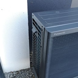 PROENERGY KAROL MIŚKIEWICZ - Zewnętrzna jednostka szarej pompy ciepła Panasonic, umieszczona na betonowych bloczkach, stoi na żwirowym podłożu przy białej i grafitowej ścianie budynku.