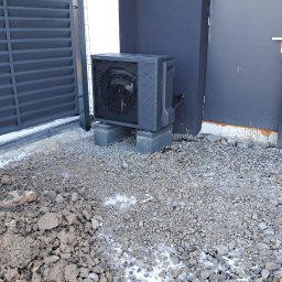 PROENERGY KAROL MIŚKIEWICZ - Czarna pompa ciepła typu split, ustawiona na betonowych bloczkach przy ścianie budynku z szarą elewacją, widoczna brama posesji, teren wokół wysypany drobnym kamieniem, miejscami resztki śniegu...