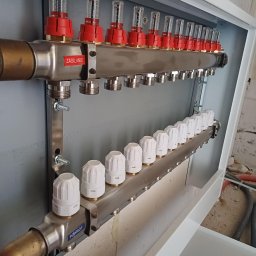 ProEnergy - Rozdzielacz ogrzewania podłogowego z przepływomierzami i zaworami termostatycznymi, zamontowany w szafce instalacyjnej, widoczne napisy 'ZASILANIE' i 'POWRÓT'.