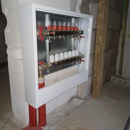 ProEnergy - Rozdzielacz ogrzewania podłogowego w białej obudowie, z czerwonymi zaworami i rurami, zamontowany na surowej ścianie z betonu komórkowego w trakcie prac wykończeniowych.