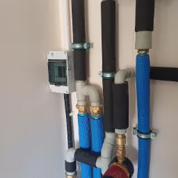 Szczegółowe ujęcie instalacji grzewczej z rurami w niebieskiej izolacji, czarnymi rurami z otuliną i pompą obiegową Grundfos Comfort PM, zamontowanej na jasnej ścianie obok skrzynki elektrycznej.