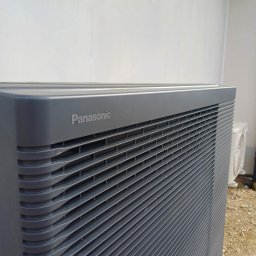 PROENERGY KAROL MIŚKIEWICZ - Szara jednostka zewnętrzna pompy ciepła Panasonic z widocznymi lamelami wentylacyjnymi i logo, na tle białej ściany i żwirowego podłoża, z drugą jednostką w tle.