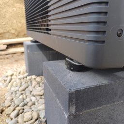 PROENERGY KAROL MIŚKIEWICZ - Szara jednostka zewnętrzna urządzenia grzewczego, prawdopodobnie pompy ciepła, ustawiona na dwóch betonowych bloczkach na żwirowym podłożu. Widoczny fragment elewacji budynku w tle.