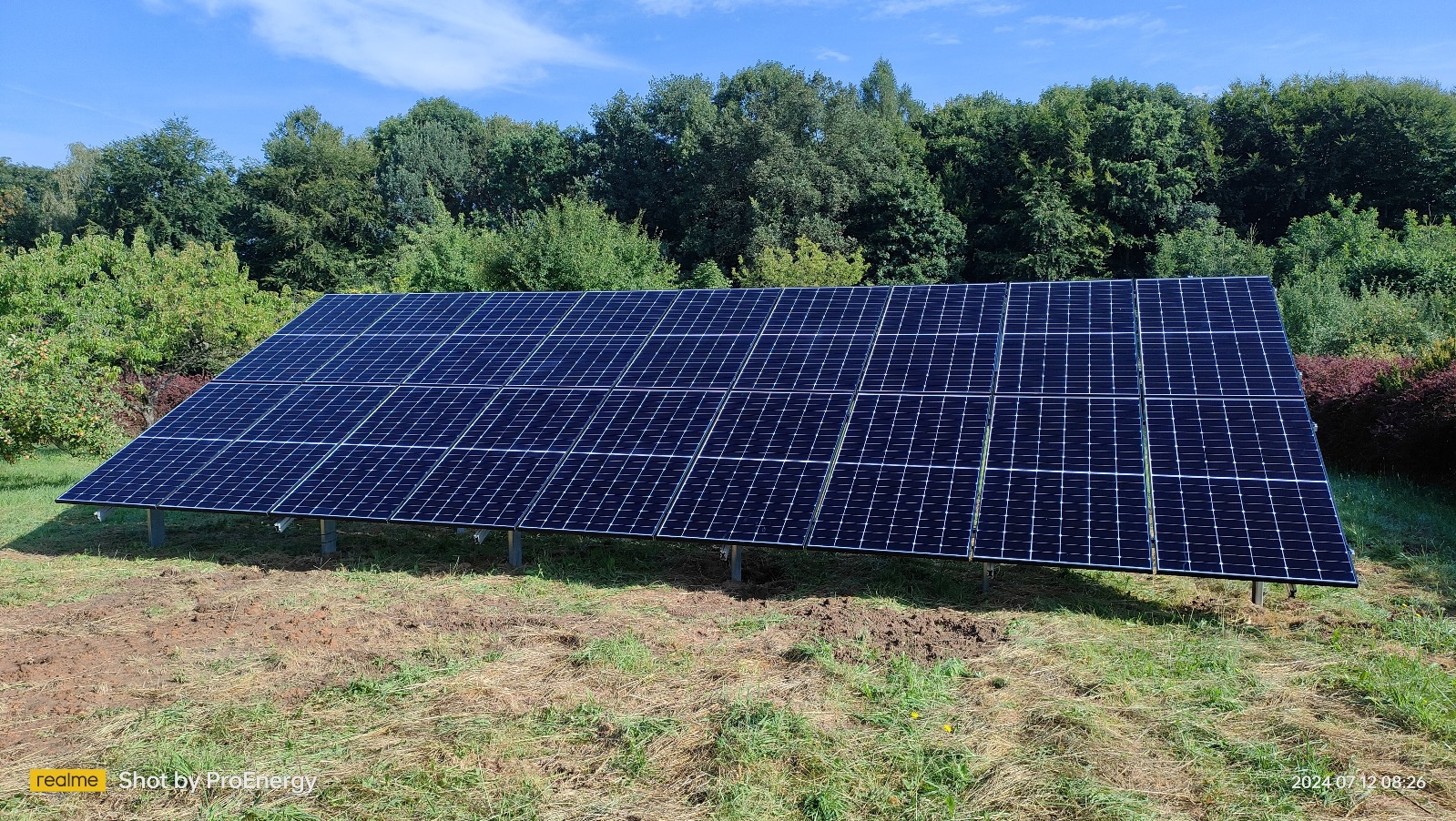 Panel fotowoltaiczny ustawiony na metalowych wspornikach, widoczny na tle zieleni drzew i błękitnego nieba, z ziemią częściowo pokrytą trawą i śladami prac ziemnych.