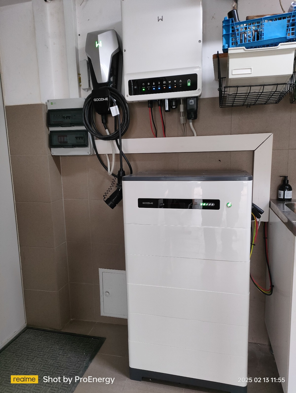 Biały magazyn energii GoodWe z podłączonymi kablami i ładowarką, zamontowany na ścianie z beżowych płytek, widoczne kontrolki stanu na urządzeniu.