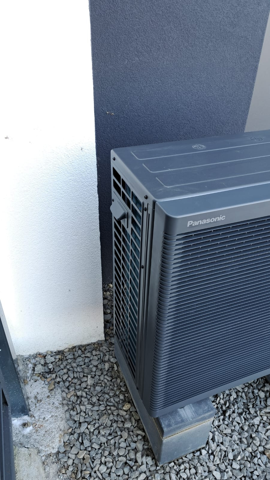 Zewnętrzna jednostka szarej pompy ciepła Panasonic, umieszczona na betonowych bloczkach, stoi na żwirowym podłożu przy białej i grafitowej ścianie budynku.