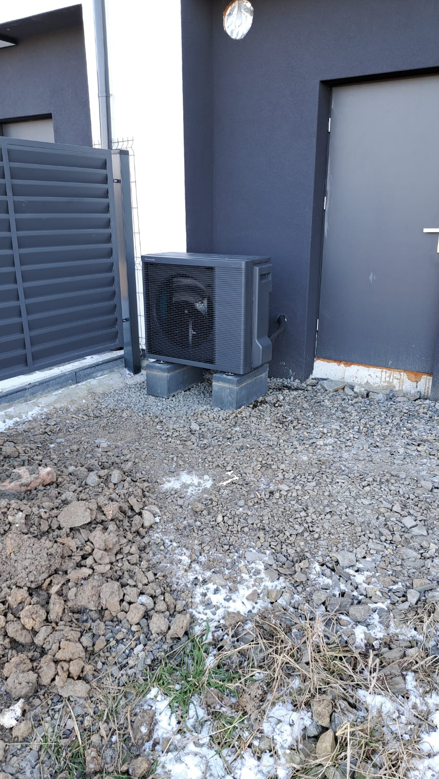 Czarna pompa ciepła typu split, ustawiona na betonowych bloczkach przy ścianie budynku z szarą elewacją, widoczna brama posesji, teren wokół wysypany drobnym kamieniem, miejscami resztki śniegu...