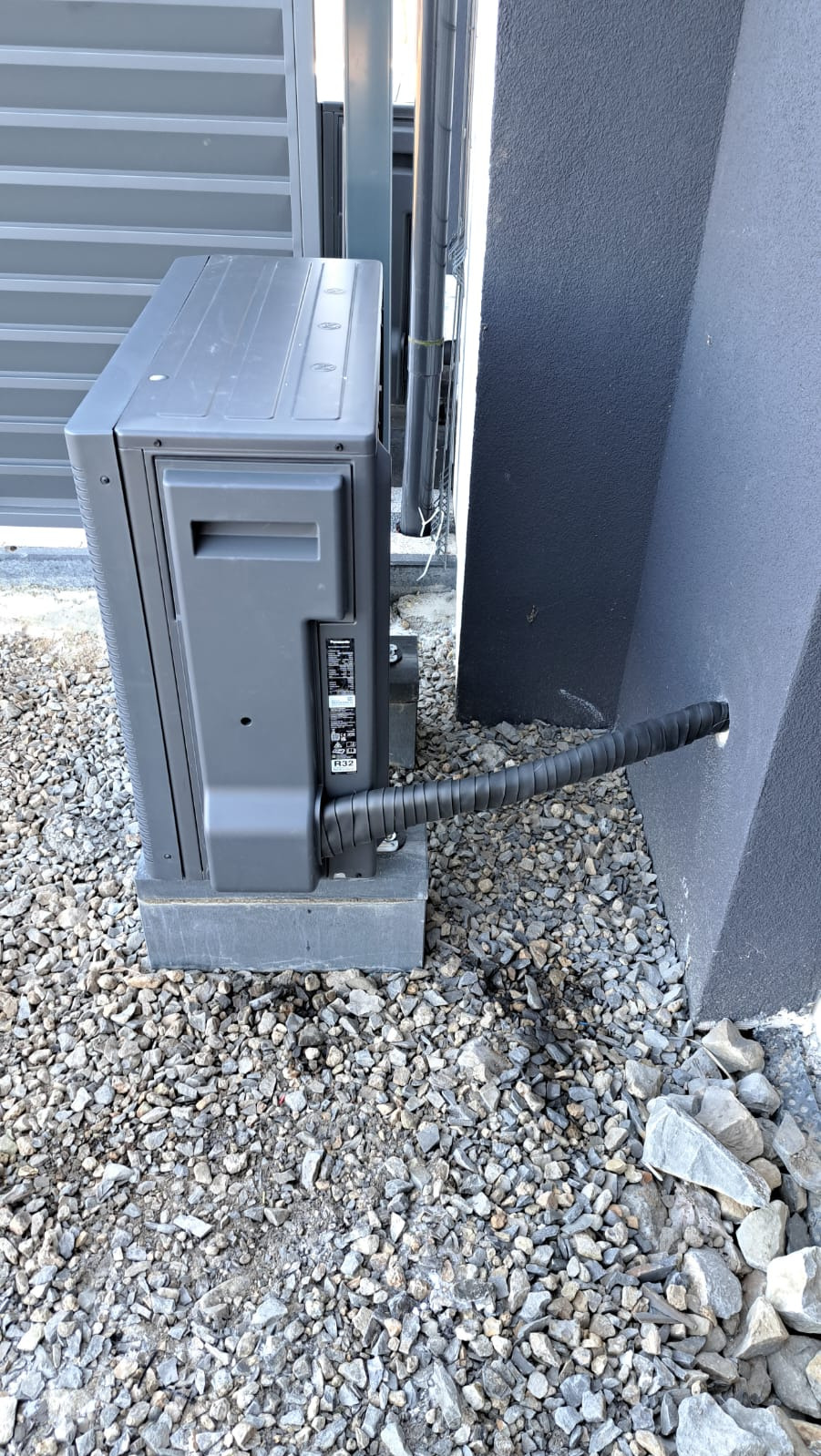 Szara pompa ciepła Panasonic R32 zamontowana na betonowym fundamencie, z widocznym przewodem w peszlu wchodzącym w ścianę budynku, otoczona drobnym szarym kamieniem.